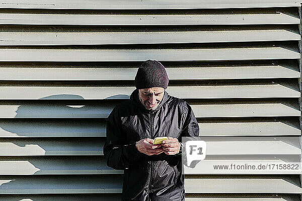 Mature man in knit hat using smart phone while leaning on shutter
