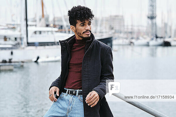 Fashionable man wearing jacket while leaning on railing at harbor