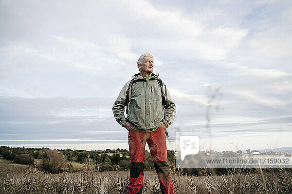 Älterer männlicher Wanderer mit den Händen in den Taschen  der auf einem landwirtschaftlichen Feld gegen den bewölkten Himmel auf dem Land steht