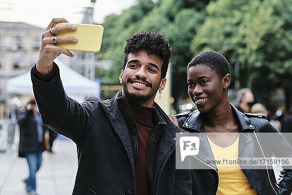 Smiling man taking selfie with friend through mobile phone while standing in city