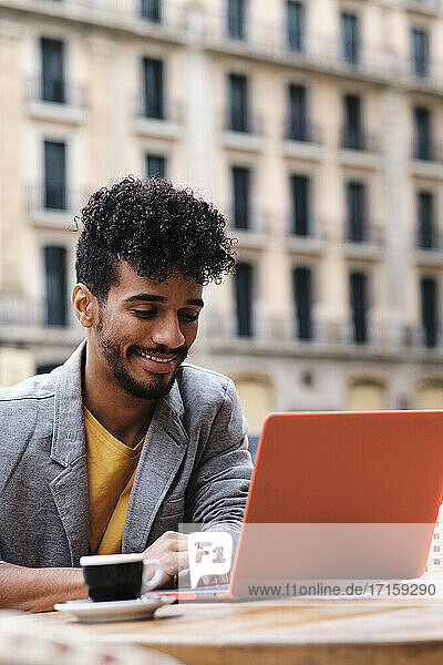 Fashionable man smiling while working on laptop sitting at side walk cafe