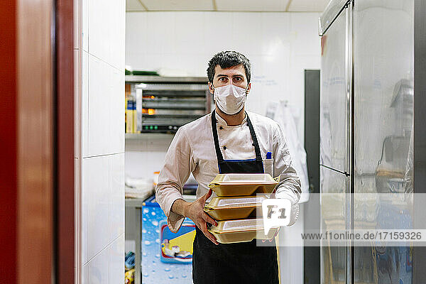 Junger Koch in Schürze mit Lebensmittelbehältern bei der Arbeit in einem Restaurant während einer Pandemie