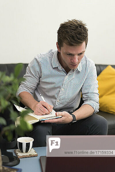 Businessman writing in book while working with laptop at home office