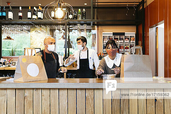 Männlicher und weiblicher Besitzer mit Koch im Gespräch bei der Zubereitung von Speisen zum Mitnehmen in einer Bar während COVID-19