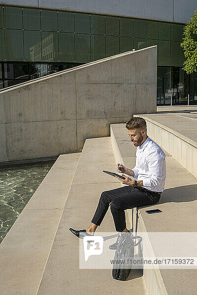 Businessman using digital tablet while sitting on steps by briefcase