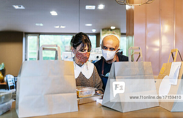 Besitzerin mit männlichem Kollegen bei der Zubereitung von Speisen zum Mitnehmen an der Theke während einer Pandemie