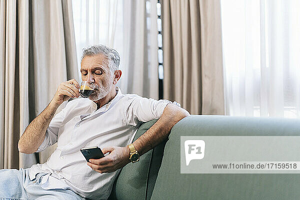 Mature man having coffee using smart phone while sitting on sofa in hotel room