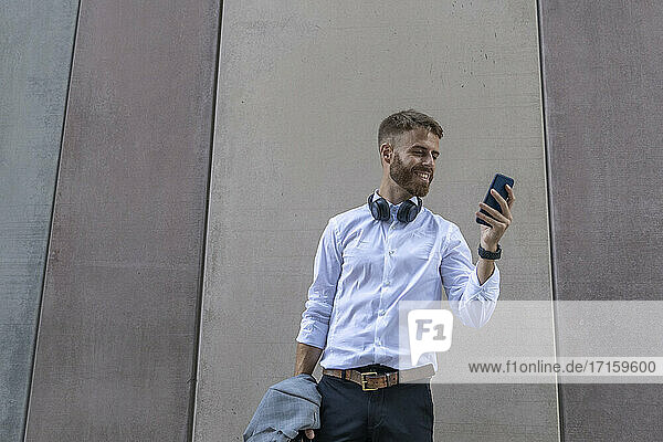 Smiling businessman using mobile phone while standing against wall