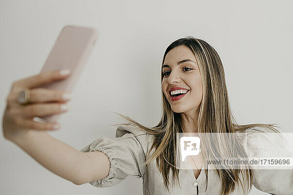 Smiling beautiful woman taking selfie through smart phone against white background