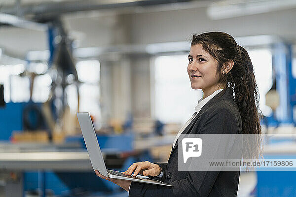 Smiling businesswoman using laptop while standing at industry