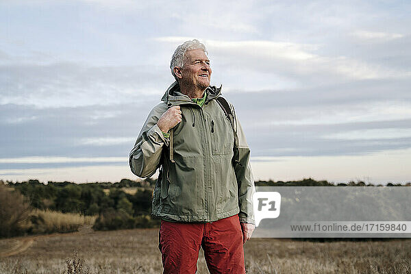 Lächelnder älterer männlicher Forscher auf einem landwirtschaftlichen Feld vor bewölktem Himmel am Wochenende