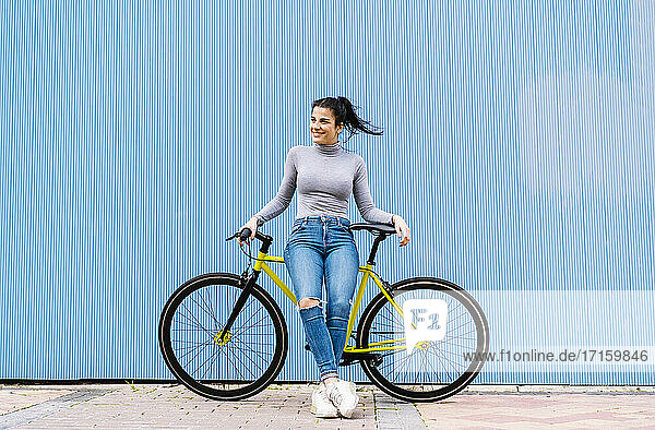 Nachdenkliche Frau lächelt  während sie auf einem Fixie-Fahrrad vor einer blauen Wand sitzt