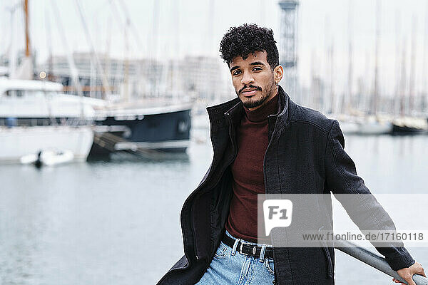 Young fashionable man wearing jacket while leaning on railing at harbor