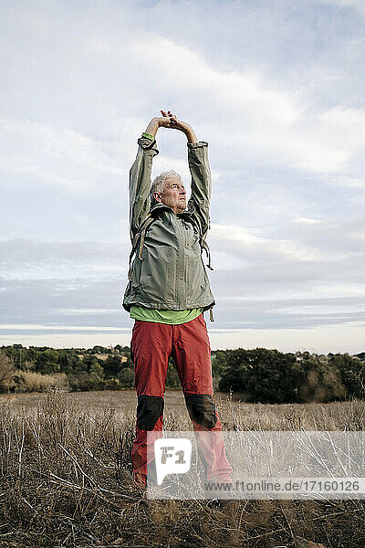 Senior male hiker stretching arms while standing on agricultural field against cloudy sky at countryside