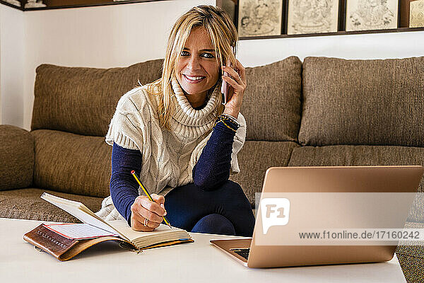 Lächelnde weibliche Therapeutin am Telefon mit Buch und Stift am Tisch am Arbeitsplatz sitzend