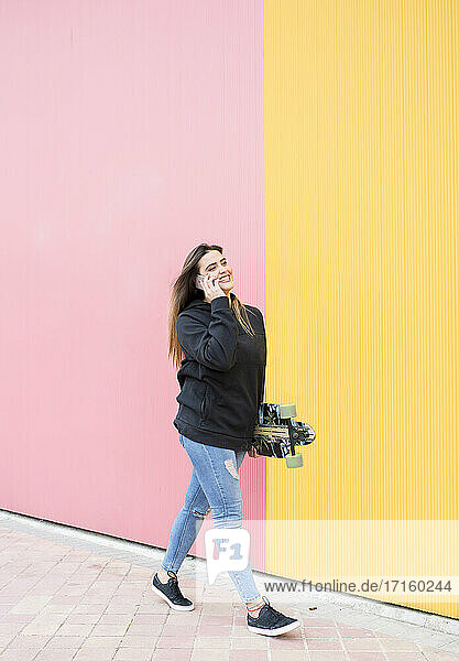 Young woman with skateboard talking on phone while walking against pink and yellow wall