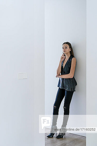 Stylish teenage girl wearing black clothing leaning on wall at home seen through hallway