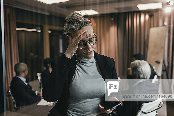 Worried mature businesswoman touching forehead while looking at smart phone against colleagues in board room
