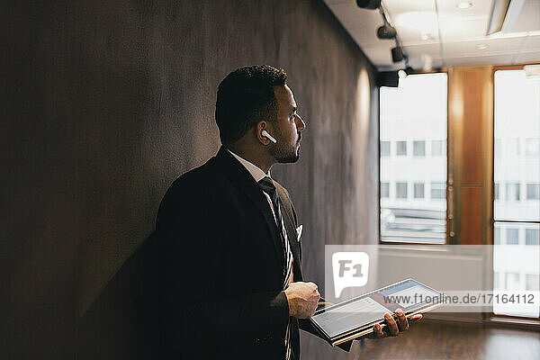 Thoughtful male professional holding digital tablet looking away while leaning against wall in board room at office