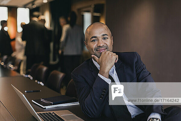Porträt eines lächelnden männlichen Fachmanns mit Glatze  der mit der Hand am Kinn am Konferenztisch im Sitzungssaal sitzt