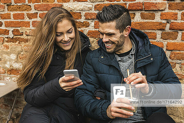 Smiling woman using smart phone while man with juice glass sitting by against wall