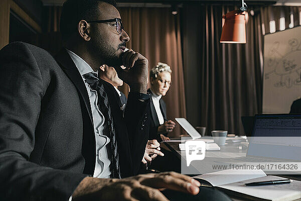 Geschäftsmann schaut weg  während er mit der Hand auf dem Kinn von Kollegen im Sitzungssaal während der Sitzung im Büro sitzt