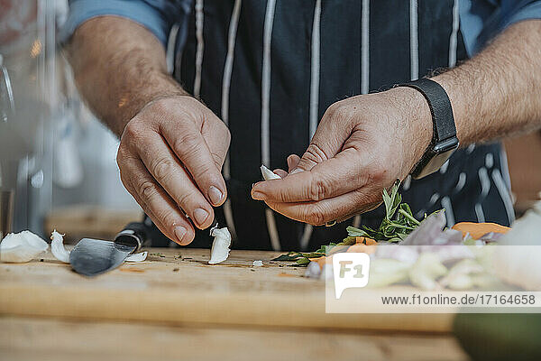 Male chef wearing apron peeling garlic while doing preparation standing in kitchen