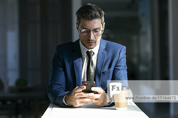 Unternehmer mit Brille  der ein Mobiltelefon benutzt  während er in der Cafeteria im Büro sitzt