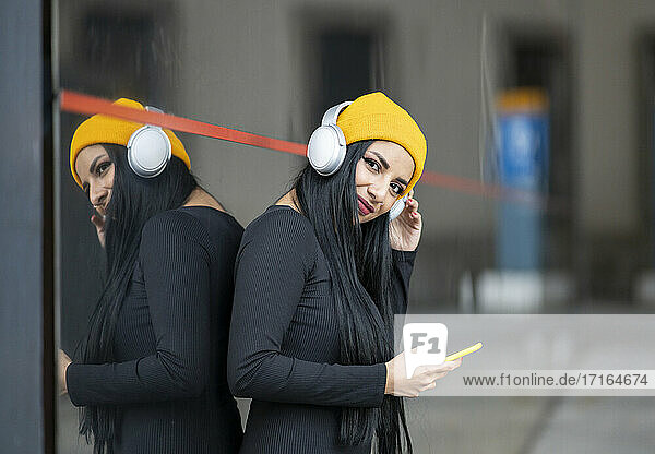 Young woman with headphones holding smart phone against glass wall