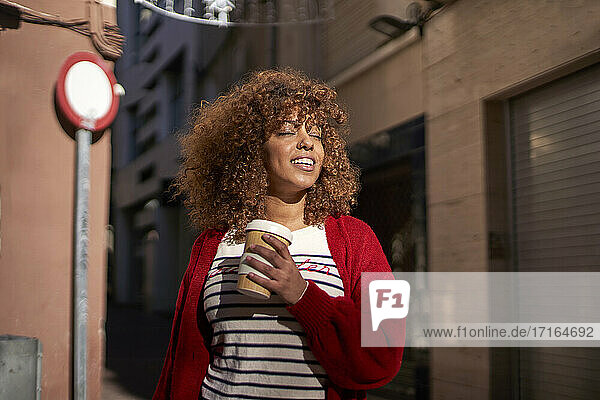 Beautiful woman with disposable coffee cup smiling while standing against street
