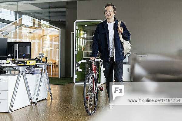 Lächelnder Geschäftsmann mit Tasche und Fahrrad  der nach der Arbeit aus dem Büro geht
