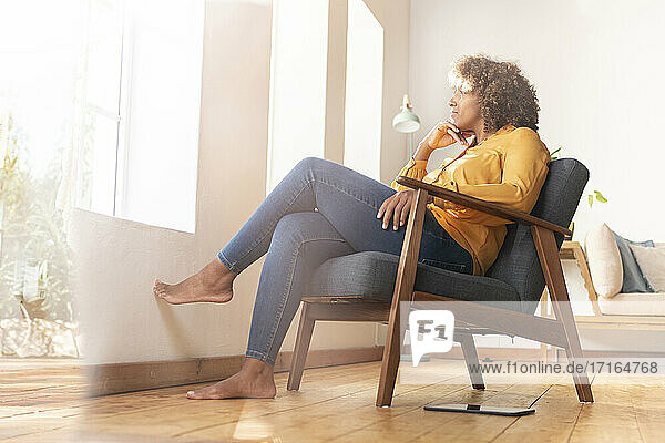 Nachdenkliche Frau mit Hand am Kinn  die durch das Fenster schaut  während sie zu Hause auf einem Sessel sitzt