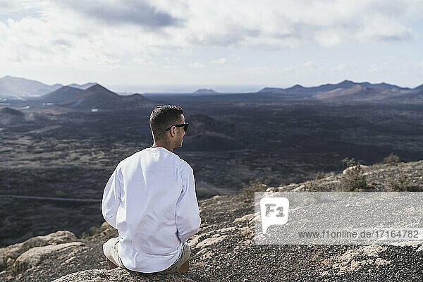 Male tourist sitting on rock while looking at view El Cuervo Volcano on sunny day  Lanzarote  Spain