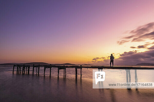 Silhouette einer Frau  die in der Morgendämmerung allein auf einem Steg an der Küste steht