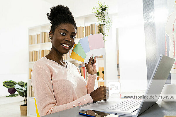Smiling fashion stylist with laptop showing colorful fabric swatch while sitting at home office