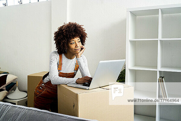 Afro-Frau mit Hand am Kinn träumt  während sie am Laptop in einer neuen Loft-Wohnung steht