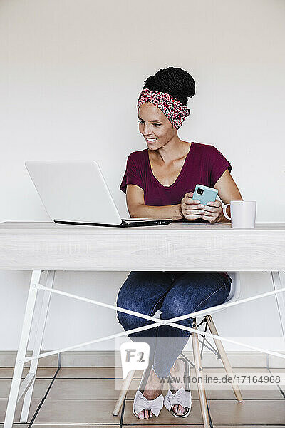 Geschäftsfrau  die einen Laptop benutzt  während sie ein Smartphone auf dem Tisch zu Hause hält