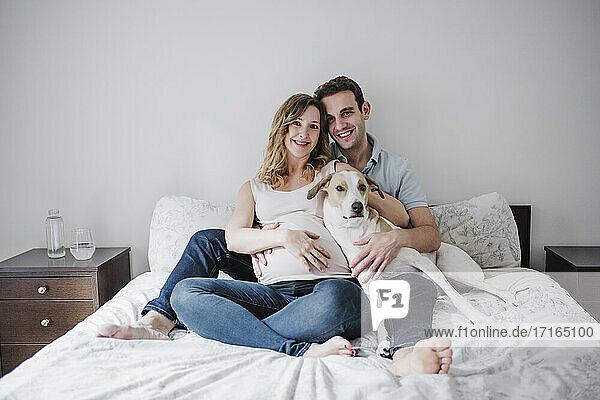 Smiling couple with dog relaxing on bed against wall at home