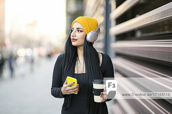 Thoughtful young woman with reusable cup listening music on smart phone while looking away at street