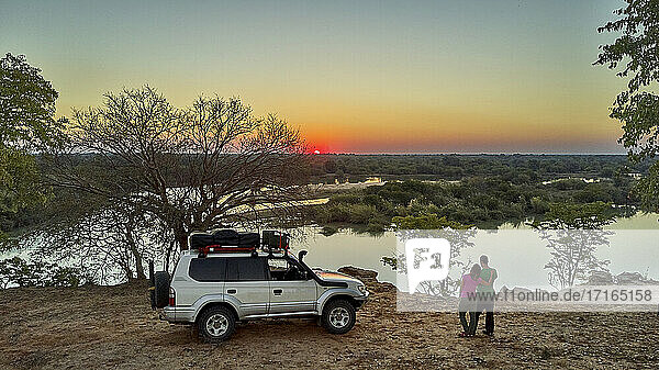 Luftaufnahme eines Paares mit ihrem Jeep  das den Sonnenuntergang vor dem Fluss genießt  Cunene-Flussgebiet  Angola