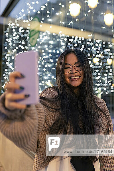 Cheerful young woman taking selfie through smart phone against illuminated glass wall