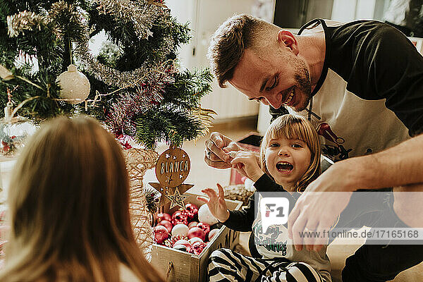 Playful daughter sitting cross-legged with family by Christmas Tree at home