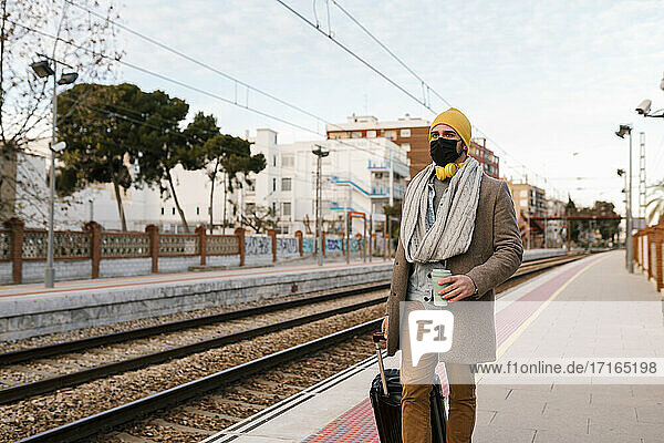 Man wearing protective mask holding coffee cup and luggage while walking on station