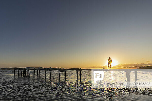 Silhouette einer Frau  die bei Sonnenaufgang allein auf einem Steg an der Küste steht
