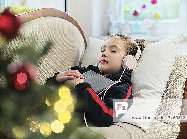 Entspanntes Mädchen  das auf dem Sofa im Wohnzimmer liegend Musik auf einem digitalen Tablet hört  während es Weihnachten feiert