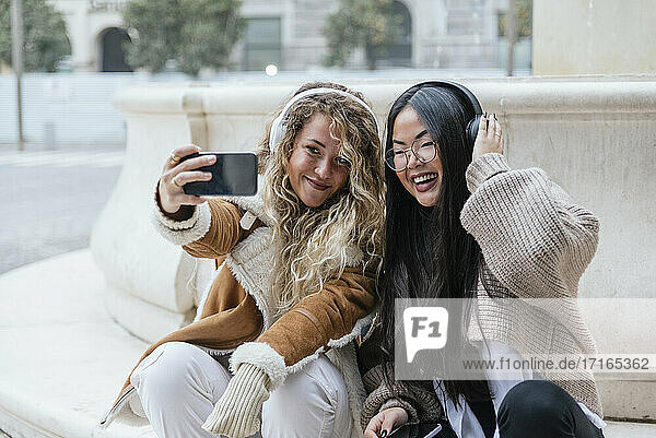 Cheerful female friends taking selfie through smart phone while listening music over headphones