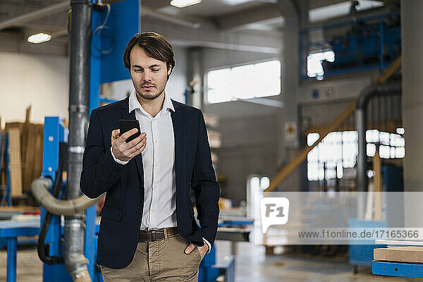 Young businessman using mobile phone while standing with hands in pockets at industry