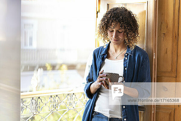 Woman looking at coffee mug while leaning on window at home