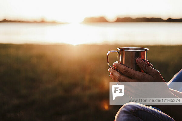 Hände einer mittleren erwachsenen Frau  die ein Getränk gegen einen See bei Sonnenuntergang hält