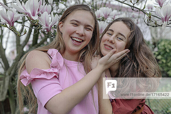 Cheerful female friends embracing against magnolia tree in public park